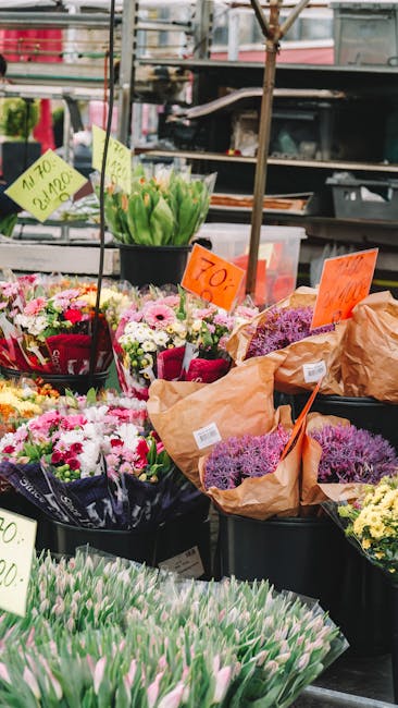 A vibrant display of fresh flowers at a flower market stall, with bouquets featuring pink, purple, and white blooms such as tulips, daisies, and chrysanthemums. The flowers are arranged in black bucke