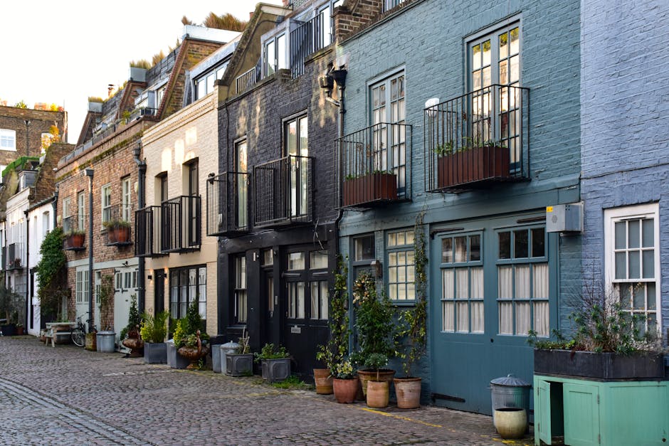 A row of charming, narrow townhouses on a cobblestone street, with each building featuring distinct pastel and neutral-colored facades. Several homes have black or blue-painted doors and large windows