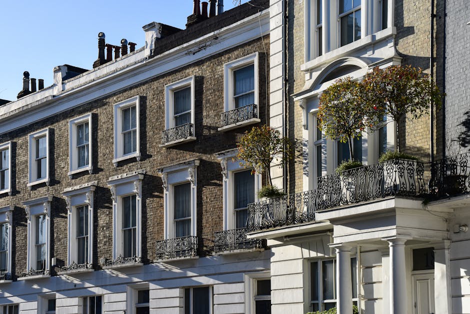A row of elegant English townhouses with brick facades and white window frames, featuring ornate black wrought iron balcony railings. One balcony, decorated with two small, well-maintained potted tree