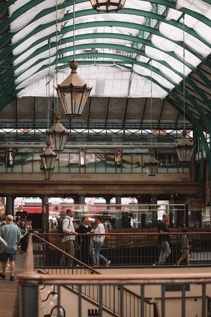 The image depicts an indoor market hall with a high, arched glass ceiling supported by green metal beams. Inside, several ornate, vintage-style lanterns hang from the ceiling, illuminating the space w