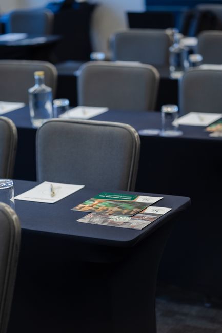 A conference room set up with several dark tables covered in black tablecloths, each table featuring a neatly arranged white notepad and pen, a clear glass water bottle, and a glass. The room has a pr