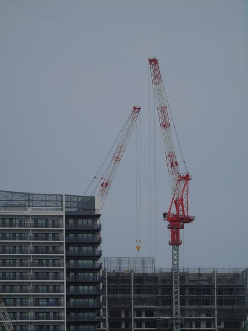 The image shows a construction site with two tall red and white cranes against a cloudy, overcast sky. One crane is slightly taller and extending diagonally to the left, while the other is positioned 
