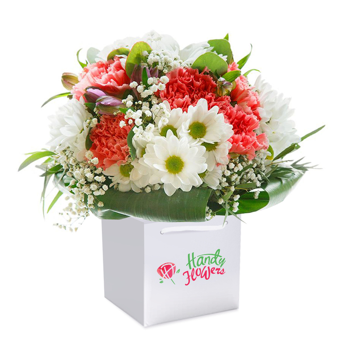 Presented upright in a minimalist white gift bag, this meticulously composed bouquet brings a touch of Covent Garden's perennial charm indoors. The starring blossoms-soft coral and rich crimson carnations-invite attention with their lavish, ruffled petals, suggesting a blend of drama and romance well-suited to the energetic district. Accentuating the bouquet's allure, crisp white daisies with lively lime-green centers peep through each cluster, reminiscent of the fresh blooms lining the local market arcade. Gentle sprays of pure white baby's breath skim the design, adding a sense of airiness and poetic calm, while nestled on the left edge, a handful of tightly closed, dusky purple-hued buds add dimension and subtle intrigue. Deep, lush greenery, including broad, gleaming leaves, cradle the arrangement in a protective, structured embrace, echoing the leafy squares of central London. The overall round, voluptuous silhouette signals freshness and expert arranging-every detail considered for a seamless finish. The white bag with its satin ribbon handles marks this as a ready-to-deliver gift, perfect for marking an opening night, birthday, or an affectionate message to someone in Covent Garden. The professionally shot photo, pure and uncluttered, directs all focus to the bouquet's artful composition and the quiet pride local florists take in their craft.
