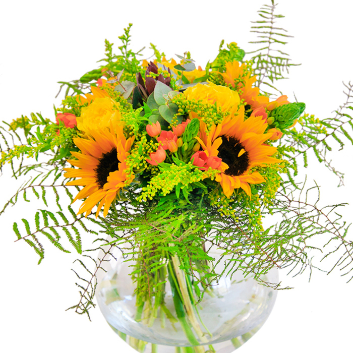 This bouquet's first impression is a rush of joy, as if Covent Garden sunshine had been gathered and arranged in a glass embrace. Large, beaming sunflowers-each crowned with tawny centers-stand tall among plush yellow roses, whose layered petals ripple outward like golden silk. Deep green leaves and trailing, lacy vines tumble from the arrangement, echoing the exuberance of the flower stalls along Floral Street. Shining red Hypericum berries glint like scattered jewels, their bright skins a playful foil to the matte repose of petals. Wisps of fern and delicate fillers dance around the edges, adding lightness and a sense of movement; slender eucalyptus leaves bring a hint of blue, subtly cooling the composition. Each stem sits revealed within the crystal-clear spherical vase, enriched by clean water-a signature of Covent Garden Florist's meticulous delivery. Up close, there's a gentle, energizing fragrance-roses, greenery, and a hint of autumn on the air. This bouquet promises to light up celebrations in the heart of the city, infusing vibrant happiness into every Covent Garden home or workspace it adorns.
